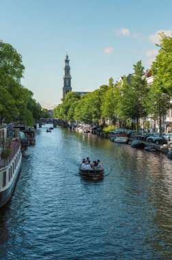 Amsterdam, Hollanda - 14 Temmuz 2025: Tarihi binalar ve botlar sıcak bir günbatımı gökyüzü altında sakin kanal sularına yansıdı.