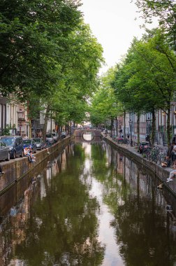 Amsterdam, Hollanda - 14 Temmuz 2025: Tarihi binalar ve botlar sıcak bir günbatımı gökyüzü altında sakin kanal sularına yansıdı.