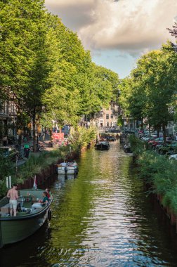 Amsterdam, Hollanda - 14 Temmuz 2025: Tarihi binalar ve botlar sıcak bir günbatımı gökyüzü altında sakin kanal sularına yansıdı.