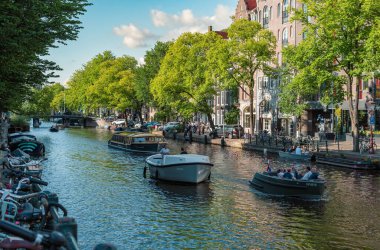 Amsterdam, Hollanda - 14 Temmuz 2025: Tarihi binalar ve botlar sıcak bir günbatımı gökyüzü altında sakin kanal sularına yansıdı.