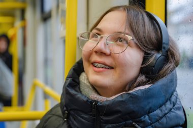 Yağmurlu bir günde tramvayda oturmuş gülümseyen ve diş tellerini gösteren neşeli bir genç kız. Şık kulaklıklar ve günlük kıyafetler giyiyor. Doğal samimi an, modern şehir yaşam tarzı..