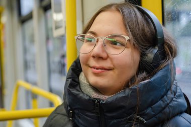 Yağmurlu bir günde tramvayda oturmuş gülümseyen ve diş tellerini gösteren neşeli bir genç kız. Şık kulaklıklar ve günlük kıyafetler giyiyor. Doğal samimi an, modern şehir yaşam tarzı..