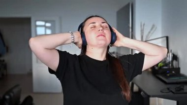 Kulaklıkla müzik dinlerken oturma odasında dans eden mutlu genç bir kadın. Rahat ev atmosferi, neşeli ruh hali, günlük kıyafet, neşeli hareket, parlak iç mekan ve pozitif enerji.