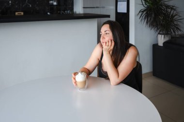 Sevimli, hafif uykulu genç bir kadın sıcak bir kapuçinonun keyfini çıkarıyor. Parlak, rahat bir mutfakta. Basit sabah rutini, rahat atmosfer, günlük yaşam tarzı, günün sakin başlangıcı..