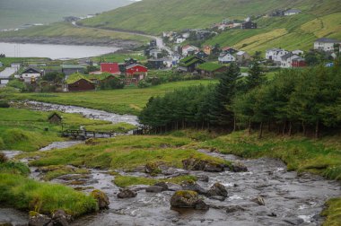 Faroe Adaları 'ndaki dramatik yeşil uçurumlar, kayalık sahil şeridi ve bulutlu kuzey havası altında okyanus dalgaları olan köyün manzarası. Sağlam, el değmemiş doğa.