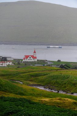 Faroe Adaları 'ndaki dramatik yeşil uçurumlar, kayalık sahil şeridi ve bulutlu kuzey havası altında okyanus dalgaları olan köyün manzarası. Sağlam, el değmemiş doğa.
