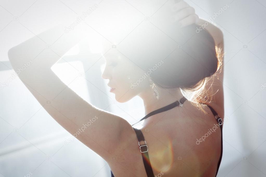 Side view portrait of the ladylike woman in the black lingerie is posing near the window surrounded by the soft light