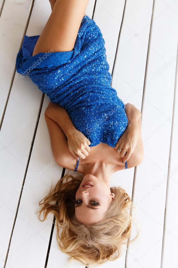 Sexy and beautiful woman in the blue evening sparkling dress with sequins is lying on the white wooden floor