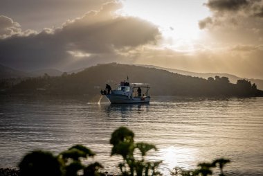 Sonbahar güneşi bulutların arasından doğuyor. Durgun denize güneş ışınları saçıyor. Küçük bir balıkçı teknesi ve balıkçı avlarını kontrol ediyor..