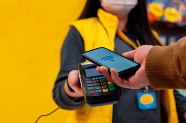 NFC teknolojisi. Erkek bir alıcı elinde kablosuz ödemesi olan bir akıllı telefon tutuyor. Virüs maskeli bir satıcının elinde ödeme terminali var.