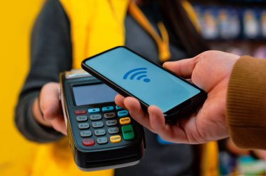Müşteri, NFC teknolojisini kullanarak akıllı telefon ile ödeme yapıyor. Ödeme terminali ve satıcının kızı ile karşılaştırıldığında,