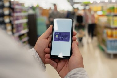 Adamın biri elinde kredi kartıyla bir marketin arka planında akıllı bir telefon tutuyor.