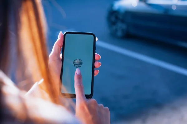 Bir kız, otoparkta elinde parmak izi sensörü olan bir akıllı telefonu arabanın arka planında tutuyor.
