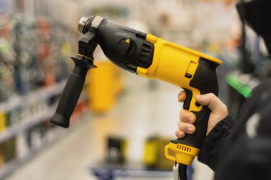 Man holds a yellow drill in his hands for repair work on the background of showcases in a hardware store
