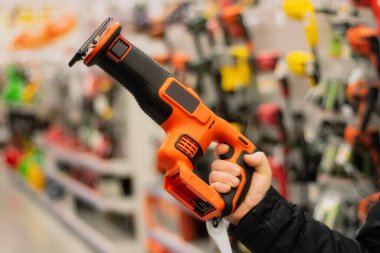 Man holds an orange reciprocating saw for repair work against the backdrop of showcases in a hardware store