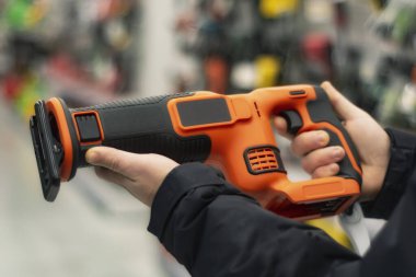 Man holds an orange reciprocating saw for repair work against the backdrop of showcases in a hardware store