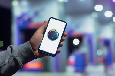 Bir adam ekranda dijital yakıt göstergesi olan bir akıllı telefonu gece benzinliğinin arka planında bir araba için tutuyor.