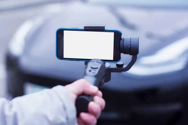 The blogger guy makes a video review of a modern car. A man holds a steadicam and a mock-up of a smartphone with a white screen in his hands