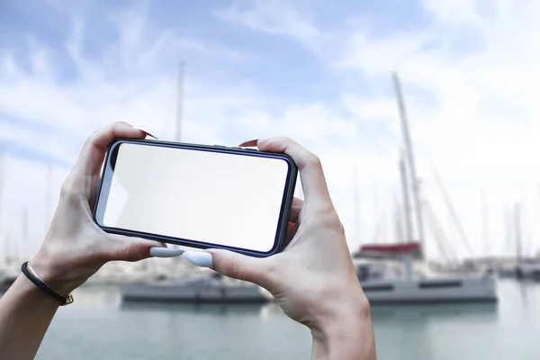 Girl holds in his hand a smartphone close-up, with a white screen against the backdrop of the sea and yachts. Mock-up Technology