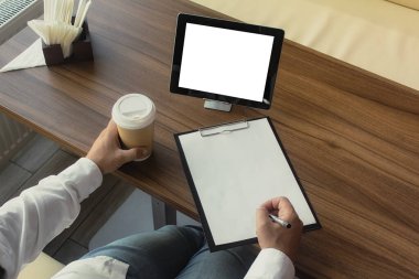 Elinde dijital tablet bulunan beyaz gömlekli bir işadamı ofiste bir sözleşme imzalıyor. Bir fincan kahve ve ahşap bir masa üzerindeki kalemli bir belge ile işyeri.