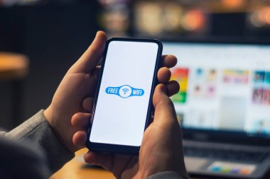 Serbest çalışan bir adam dizüstü bilgisayarının arka planında ücretsiz internet bulunan bir akıllı telefon tutuyor.