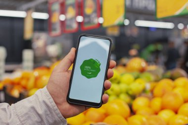 Doğal ürün. Bir adam elinde elinde bir akıllı telefon, ekranında bir etiketle süpermarketteki bir vitrinde taze yeşilliklerin arka planına karşı duruyor. Çevre dostu ve sağlıklı yiyecekler.