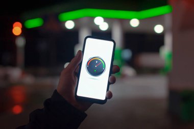 Bir adam ekranda dijital yakıt göstergesi olan bir akıllı telefonu gece benzinliğinin arka planında bir araba için tutuyor.