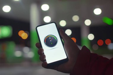 Bir adam ekranda dijital yakıt göstergesi olan bir akıllı telefonu gece benzinliğinin arka planında bir araba için tutuyor.