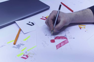 Grafik tasarımcısı bir masa üzerindeki çizim ve çizimlerin arka planına karşı bir logo tasarlar. Stüdyoda dizüstü bilgisayarla kağıda basılmış logolar.
