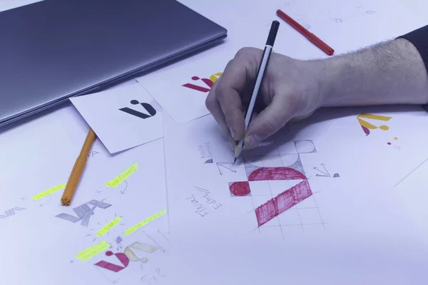 Grafik tasarımcısı bir masa üzerindeki çizim ve çizimlerin arka planına karşı bir logo tasarlar. Stüdyoda dizüstü bilgisayarla kağıda basılmış logolar.