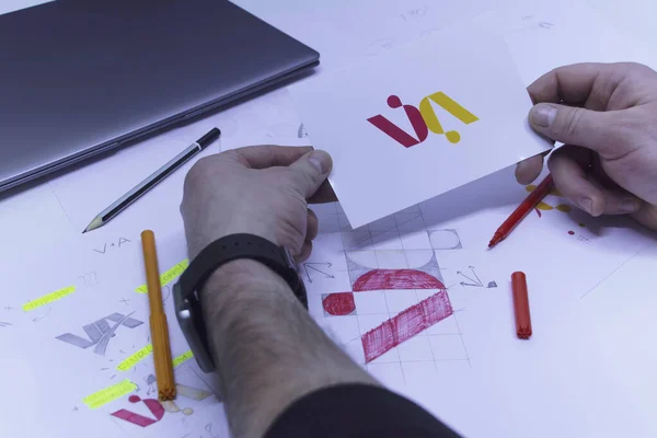 Graphic designer designs a logo against a background of sketches and drawings on a table. Printed logos on paper in a studio with a laptop