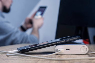 Telefon ve kablo olan bir güç bankası monitörde oturan bir adamın arka planında. Taşınabilir şarj aleti ahşap bir masada akıllı telefonu şarj eder.
