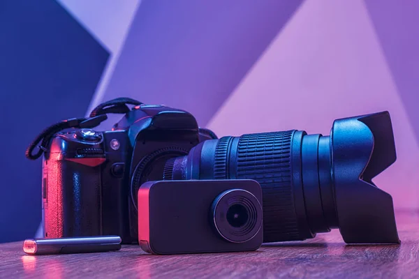 Set of equipment for photography and video filming. Photo camera with lens, action camera and usb flash drive on a wooden table in a studio with creative light