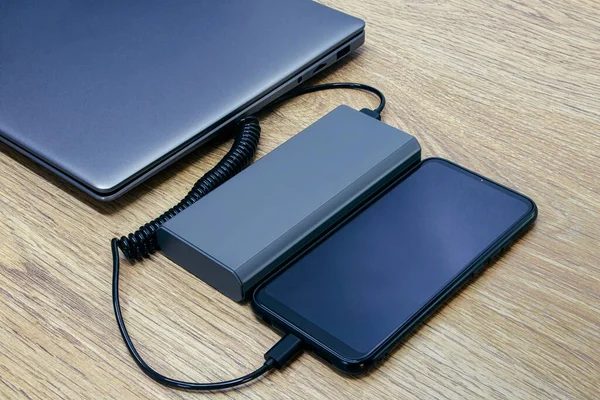 Akıllı telefon bir elektrik bankasından para alıyor. Tahta bir masada dizüstü bilgisayarı olan taşınabilir şarj aleti.