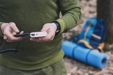Turist bir adam akıllı telefonu elinde tutuyor ve onu elektrik bankasıyla suçluyor. Sırt çantası ve ormanda turist halısı.