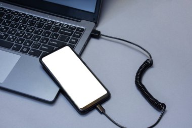 Gri bir masanın arka planında dizüstü bilgisayardan şarj edilen beyaz ekranlı bir akıllı telefonun maketi.