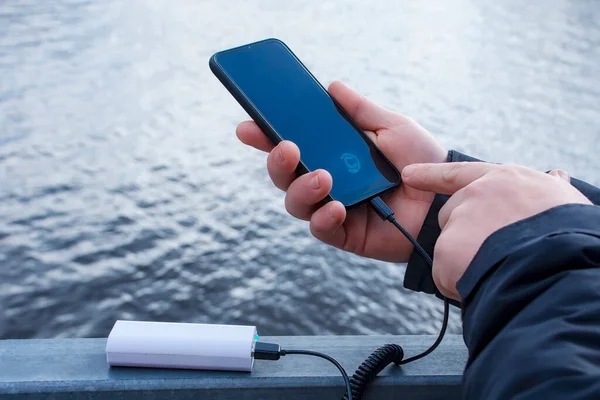 Bir adam akıllı telefonu elektrik bankasıyla şarj ediyor. Eldeki telefon gölün arka planında taşınabilir şarj cihazıyla şarj ediliyor.