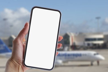Kız elinde beyaz ekranlı bir akıllı telefonun maketini tutuyor. Havaalanının arka planında bir uçak var.