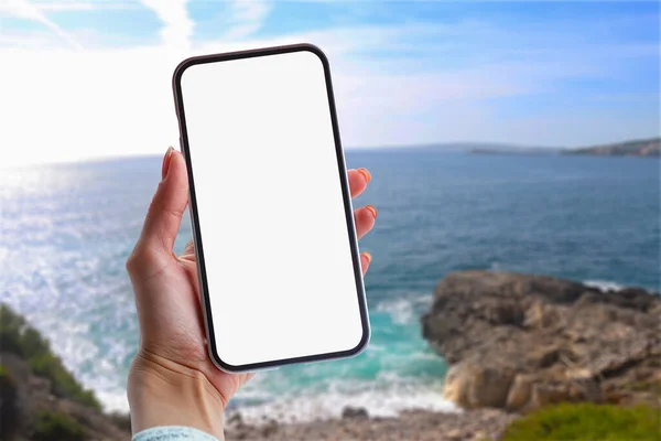 Girl holds in his hand a smartphone close-up, with a white screen on a background of sea. Mock-up Technology