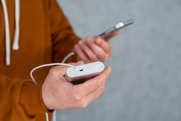 Man holds a smartphone and a portable charger in his hands on a gray background. Power Bank charges the phone