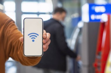 Online ödemeler. ATM ATM 'nin arka planında kablosuz ağ ikonu olan bir telefon görüntüsü. Adam elinde akıllı bir telefon tutuyor.