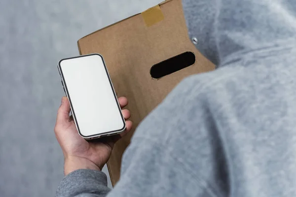 Man from a delivery service with a parcel in his hands holds a mock-up of a smartphone on a light background