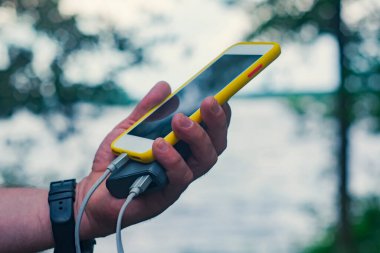 Adamın elinde akıllı telefonu olan taşınabilir bir şarj aleti var. Doğanın arka planında yeşil ve gölü olan bir adam..