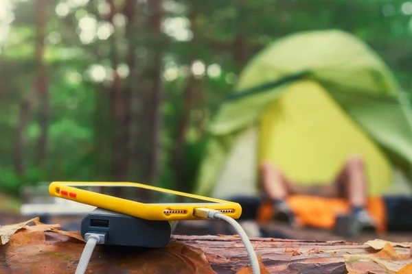 Smartphone is charged using a portable charger. Power Bank charges the phone outdoors with a backpack against the backdrop of a tent and a tourist