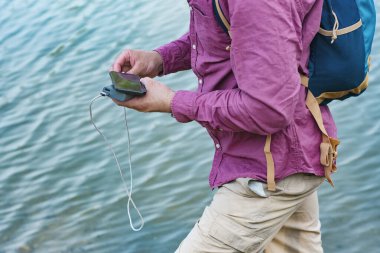 Adam akıllı telefonu taşınabilir şarj cihazıyla şarj ediyor. Parlak güneşli bir günde elinde bir güç bankası olan bir adam denizin arka planında bulutlarla