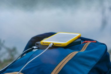 Akıllı telefon taşınabilir şarj aleti ile şarj edilir. Elektrik Bankası açık havada telefon faturasını doğa ve nehir arka planında turizm için bir sırt çantasıyla ödüyor.