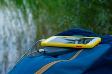 Akıllı telefon taşınabilir şarj aleti ile şarj edilir. Elektrik Bankası açık havada telefon faturasını doğa ve nehir arka planında turizm için bir sırt çantasıyla ödüyor.