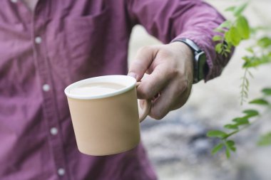 Adam, elinde kahve olan bir kupayı doğanın arka planında tutuyor.
