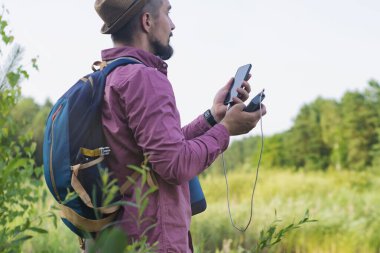 Turistin elinde taşınabilir şarj aleti olan akıllı bir telefon var. Güç bankası olan bir adam telefonu doğanın ve ormanın zeminine karşı dolduruyor.