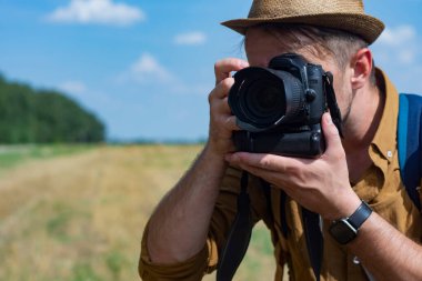 Gezgin fotoğrafçı elinde kamerayla bir tarlanın arka planına ve samanlıklara bakıyor.